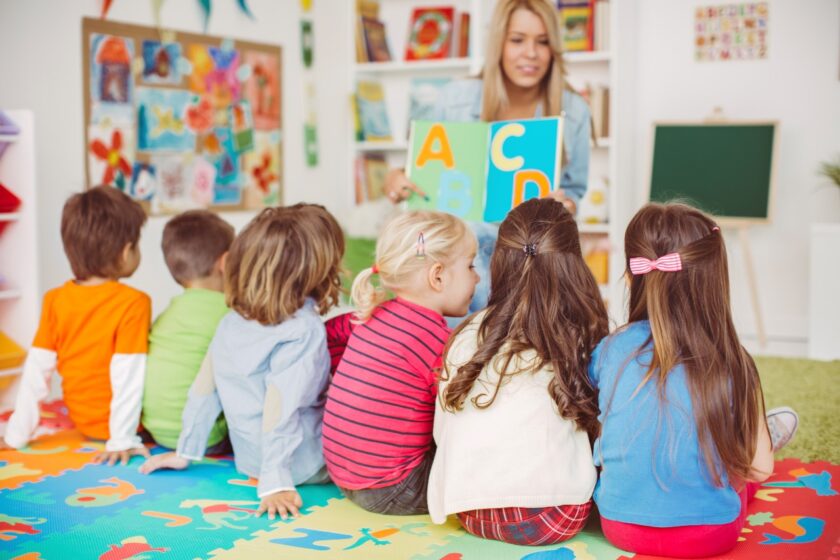 A teacher shows alphabet cards to a group of young children sitting on a colorful play mat in a classroom.